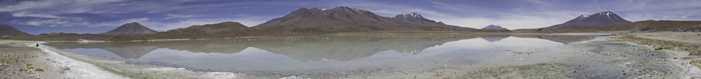 A long Altiplano lake mirroring the full Andean range, flamingos scattered across the still water