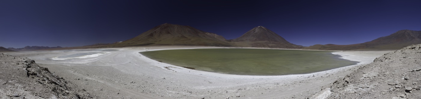 The full sweep of Laguna Verde and the Licancabur volcano across the Bolivian Altiplano, rocky shore framing the emerald water