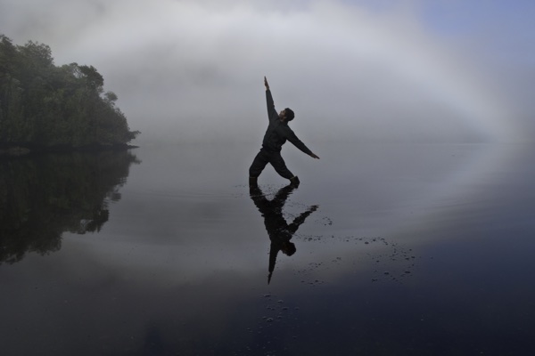 Sam reaching for a faint rainbow over a glass-still inlet in the Chilean fjords, their reflection touching the rainbow's reflection on the water
