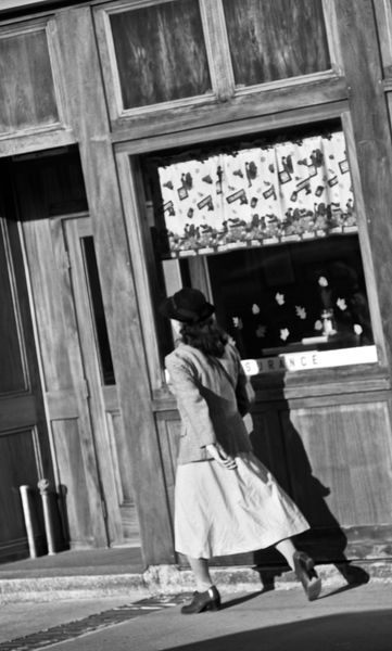 A woman in a hat and full skirt walking past an old wooden storefront, black and white